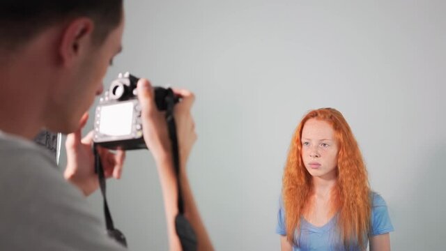 Young professional photographer taking photos of a red haired girl in a studio with white background. Rear view. Girl with lots of freckles on the face. Slow motion