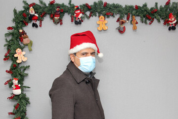 latin man with protection mask, coat and santa claus hat in christmas decoration, new normal...