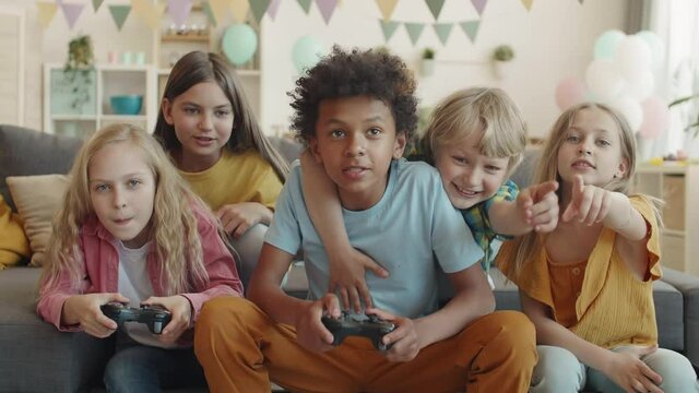 Medium POV of multiethnic school friends sitting on couch in room talking, holding game console controllers. African boy winning over Caucasian blond girl in computer game