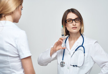 A woman nurse holds a syringe in her hand on a light background with a stethoscope and a female patient