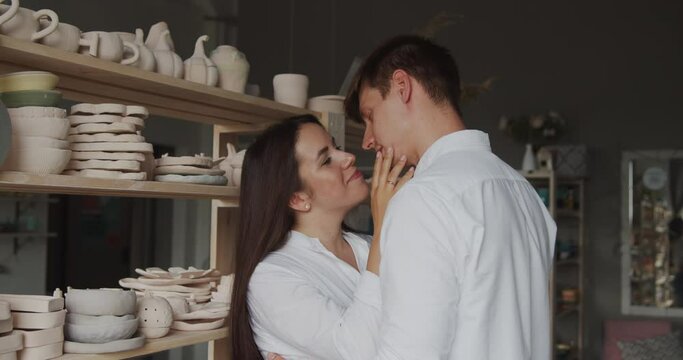Loving young couple enjoying romantic date in pottery workshop. Lovers in white shirts stay close to each other in front of the shelves with clay dishes touching each other and smiling.