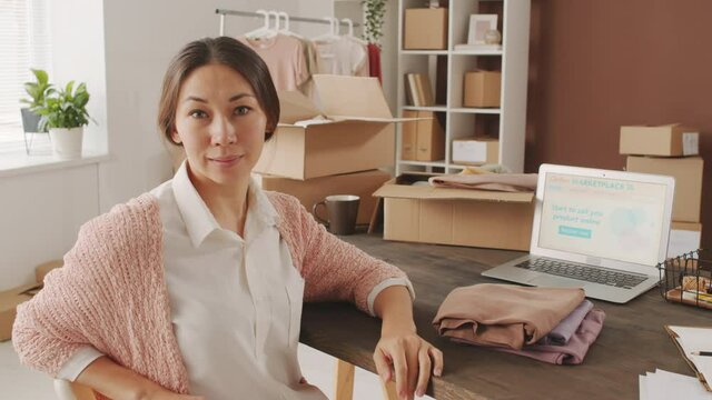 Portrait shot of happy young Asian woman smiling for camera in office of small online clothes store. Folded garments and laptop with online marketplace web page open on it are on table