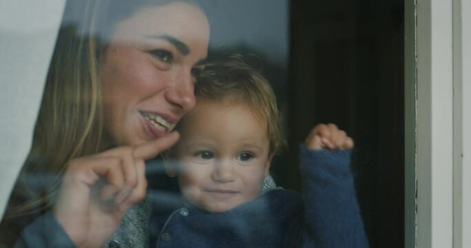 Cinematic shot of an young happy smiling mother is enjoying time together with her toddler baby boy while looking through a window in the morning. Concept of happiness ,childhood, life, motherhood.