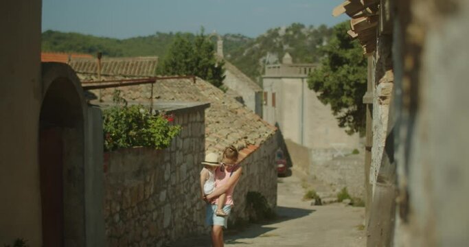 Wide shot of a young teenage girl holding her 2 years old sister in her arms walking in the streets of an old european town on a sunny day in summer