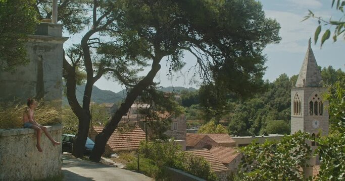 Wide shot of a young cute teenage girl sitting in the grass in her own and waving her feet, nearby a church in the middle of an empty old european town in Lastovo island Croatia on a sunny day