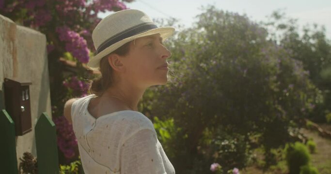 Mid shot of a woman standing standing looking around her besides a fence and a garden on a sunny day