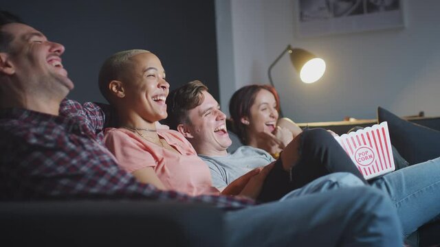 Group of friends sitting on sofa at home eating popcorn and laughing watching evening comedy TV together - shot in slow motion