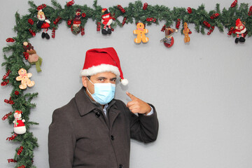 latin man with protection mask, coat and santa claus hat in christmas decoration, new normal...
