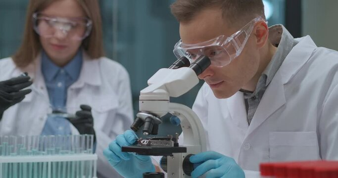 pair of scientists is working in crime lab, man is viewing evidence in microscope, woman is testing liquids in test tubes