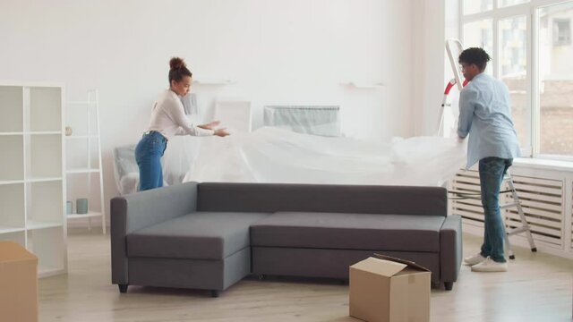 Wide shot of young diverse man and his beautiful girlfriend uncovering sofa in their new apartment and starting moving it