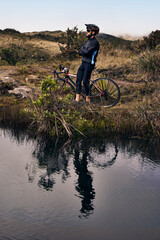 Young cyclist in black windbreaker jacket, protective helmet and glasses, taking a break in mountains and lake after riding a bike