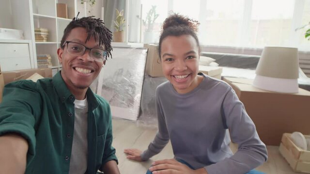 Medium shot of young African-American man and his pretty wife sitting together on floor at home and talking to friends by video link while getting ready for move