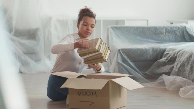 Lockdown of young beautiful mixed-race woman sitting on floor in her new apartment and unpacking her stuff while unrecognizable male loader carrying cardboard boxes