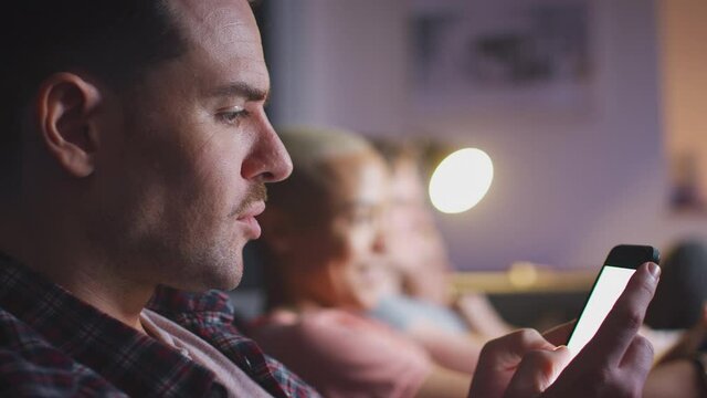 Man using mobile phone whilst friends watch TV at home in evening - shot in slow motion