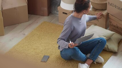 High angle view of young mixed-race woman wearing casual clothes sitting on floor at home and making list of packed stuff for move