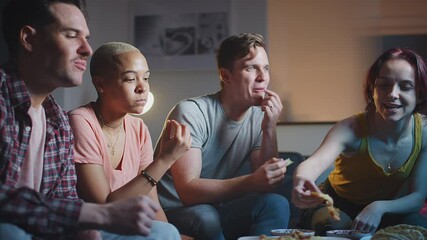 Group of friends sitting on sofa at home eating snacks and laughing watching evening comedy TV together - shot in slow motion