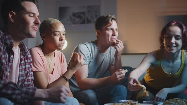 Group of friends sitting on sofa at home eating snacks and laughing watching evening comedy TV together - shot in slow motion
