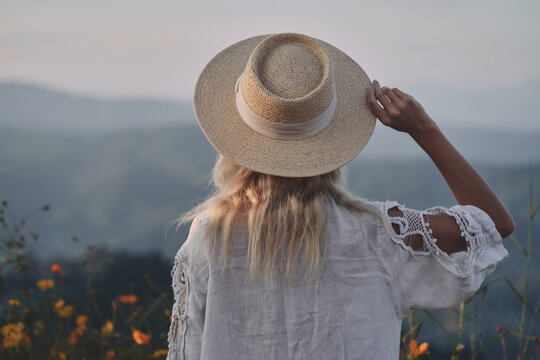 The blonde in a linen dress and straw hat walks among herbs and wildflowers on a meadow in the mountains. A woman in the Boho style, resting in the countryside, a simple village lifestyle