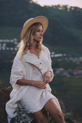 The blonde in a linen dress and straw hat sits on a viewpoint in the mountains. A woman in the Boho style, resting in the countryside, a simple village lifestyle