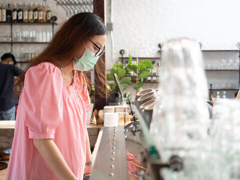 Asian Woman In Medical Face Mask Looking At Coffee Menu In Cafe. Female Buying Drinks At Cashier.  