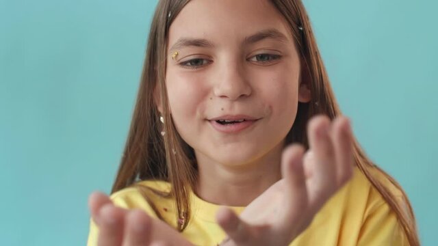 Close-up of brown-haired Caucasian school girl holding handful of sparkles, breathing in deeply, and blowing it off on camera. Female kid posing on light blue background smiling