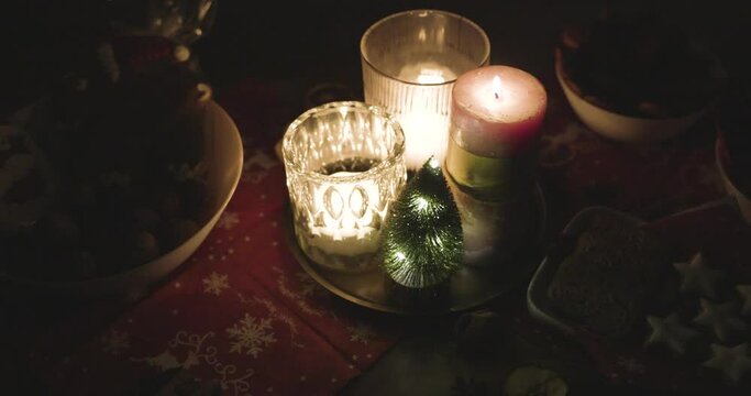 burning candles in darkness on a beautifully decorated christmas table with christmas cookies, nuts, gingerbread, dried fruits, cinnamon stars, cinnamon sticks and a christmas tree in background in 4k