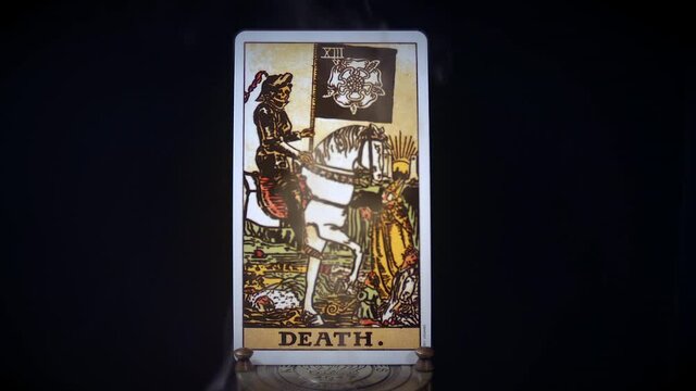 A tarot death card sits on a brass stand as smoke rises around it. The grim reaper figure is displayed on a black background. The knight sits on his white smokey horse.