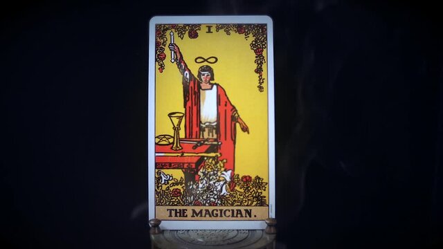 A tarot Magician card sits on a brass stand as smoke rises around it. The mystical magical figure is displayed on a black background. The robed man raises a wand.