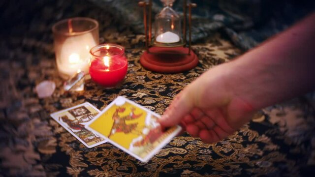 A mystical fortune teller lays out a tarot card spread. The death, fool, moon, broken heart and hangedman cards are displayed on a mysterious table adorned with candles, and hourglass and crystals.