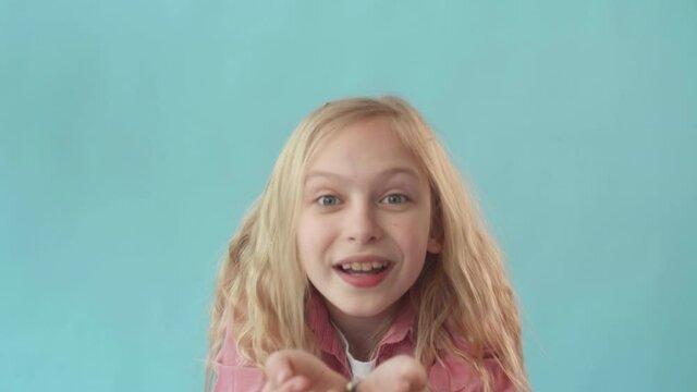 Portrait of blond-haired Caucasian school girl holding handful of sparkles, breathing in deeply, and blowing it off on camera. Female child standing in light blue background smiling