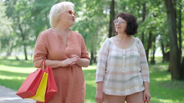 Two positive Caucasian senior women strolling in sunny spring or summer park with shopping bags and talking. Portrait of happy relaxed retirees walking outdoors and chatting. Eternal friendship.