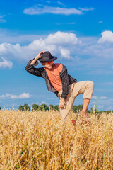 Tall handsome man with black hat and pink sunglasses standing with one leg on a brown vintage leather suitcase at golden oat field.