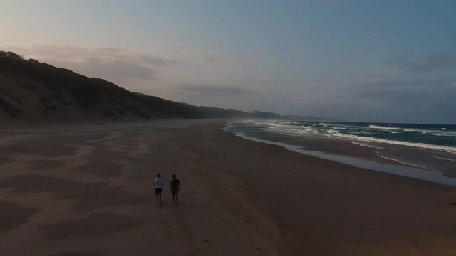 Aerial track shot of two male people walking on sandy beach beside waves from the ocean and cliffs from the mountains.
