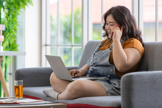 smiling fat  woman sitting on a couch talking on a smartphone and looking at a laptop computer to communicate with people in a house. Work from home concept
