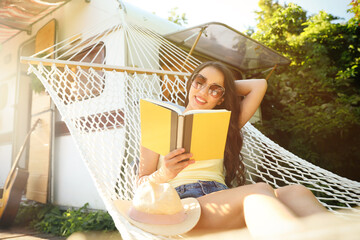 Young woman reading book in hammock near motorhome outdoors on sunny day