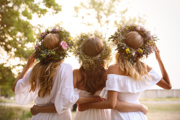 Young women wearing wreaths made of beautiful flowers outdoors on sunny day, back view