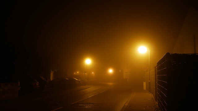 Dorfstrasse in der Nacht im dichten Nebel mit Strassenlaternen, die fahl leuchten und ein Gef&uuml;hl der Einsamkeit und des Grusels erzeugen