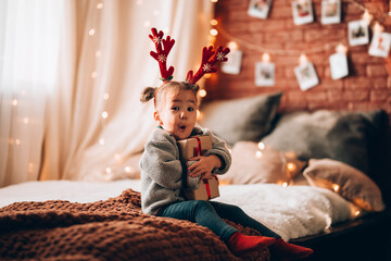 A little girl holding a large gift in a paper box on a large bed. The child has antlers on his...