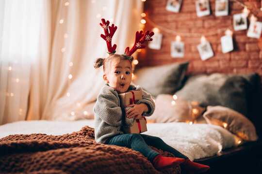A little girl holding a large gift in a paper box on a large bed. The child has antlers on his head. Christmas mood