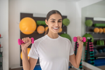 Smiling dark-haired female lifting pink dumbbells in the gym