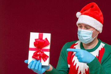 Handsome male wears Santa hat medical mask gloves showing at gift box red studio background.