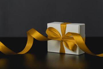 Close-up view of white gift box with elegant yellow ribbon in luxury bow on dark background and reflection. Gift box with place for text. For birthday, xmas, anniversary presents, gift post cards
