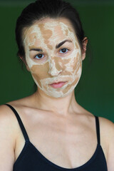 young brunette woman with a clay homemade mask on her face looks into the frame