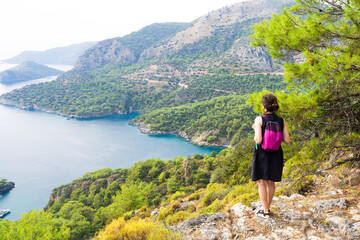 Naklejka na ściany i meble A girl on a trekking trip in the mountains, admires the beautiful natural view of the bay of the sea and the mountains of the mediterranean