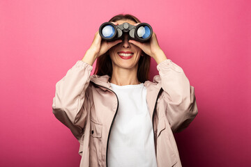 Smiling young woman in jacket looking through binoculars on pink background.