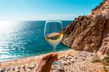 A glass of white wine in a man's hand. Wine against the backdrop of the sea coast and a sunny summer beach. Wine tasting and relaxation at the resort