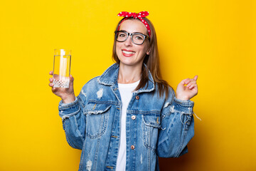 Young woman in jeans, glasses and a headband holding a glass of clean water on a yellow background