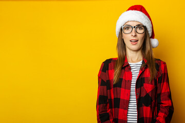 Young woman with surprised face and santa hat and red checkered shirt on yellow background