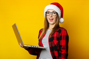 Young woman with surprised face in santa hat and red checkered shirt holding laptop on yellow background