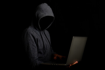 Man without a face in a hood holds a laptop on a dark background. Concept of cyber rogue, hacker....
